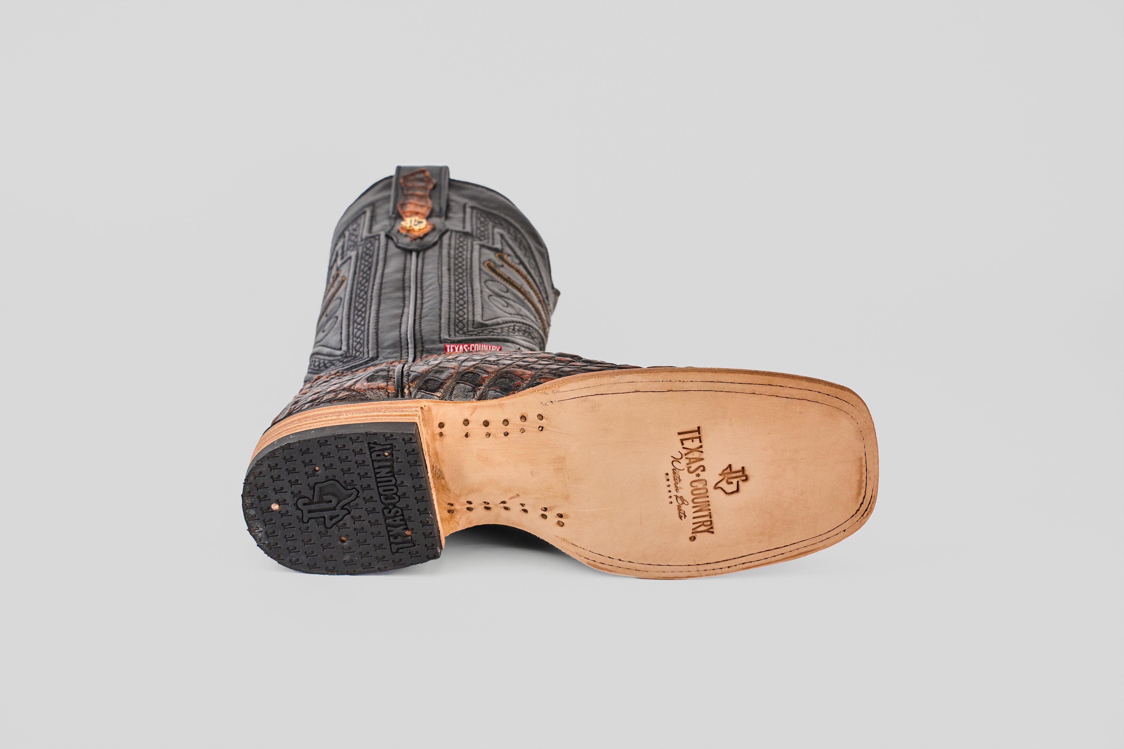 The Exotic Caiman Horn Back - Roma - Square Toe boot lies on its side, displaying a leather sole with the Texas Country logo, a wooden heel with rubber grip, and intricate upper stitching against a light gray background.