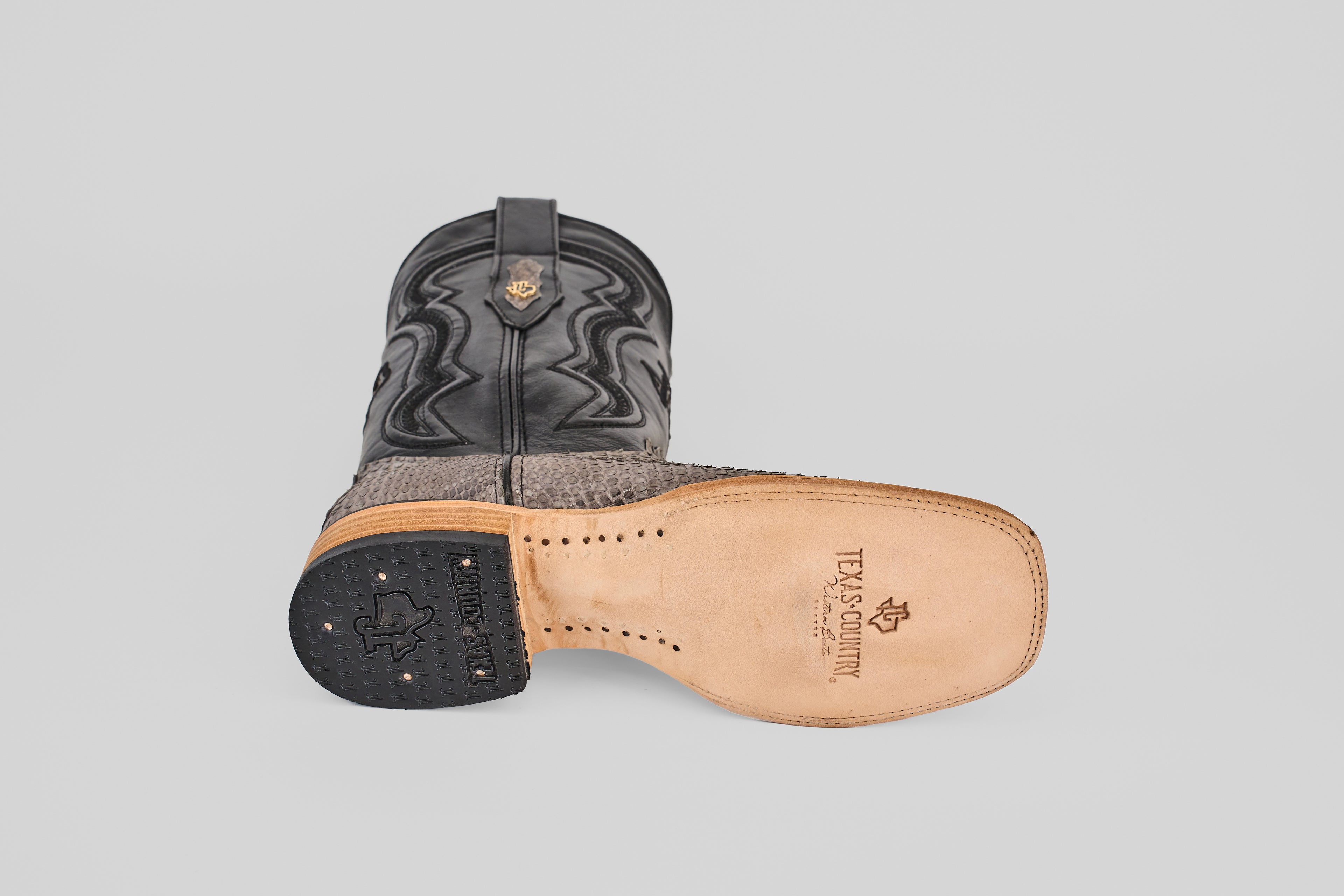 The Tanner - Matte Gray - Square Toe boots feature one upright and one on its side to display the Texas Country-branded sole, with a decorative stitched upper and textured Python Gris Nobuck lower section.
