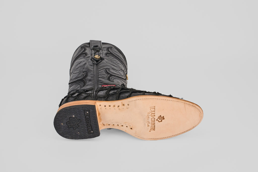 The Conor - Matte Black - Roper Toe boot is shown on its side, highlighting detailed stitching and a light tan leather sole with Texas Country branding and decorative nails—a true mark of classic western style.