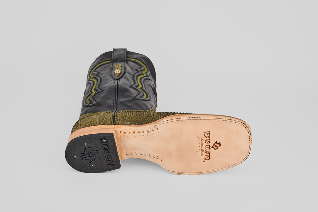 The Tanner - Green Jungle - Square Toe cowboy boot lies on its side, displaying a tan leather sole stamped with Texas Country. Its black upper is detailed with green stitching, and the lower section has a python-textured green finish.